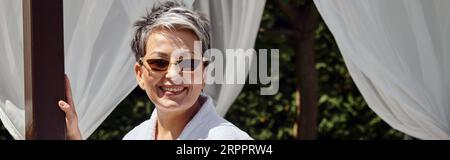 Freudige reife Frau in Sonnenbrille, die im privaten Pavillon ruht, Wellness-Retreat im Resort, Banner Stockfoto