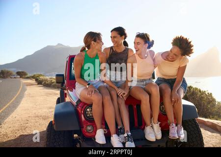 Lachende Freundinnen Sitzen Auf Der Haube Eines Open Top-Autos Auf Der Road Trip Stockfoto