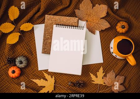 Foto von oben von einem offenen, leeren Planer, Notizbuch, einer orangefarbenen Tasse und Herbstblättern auf braunem, erdfarbenem Stofftexturhintergrund. Herbst Stockfoto