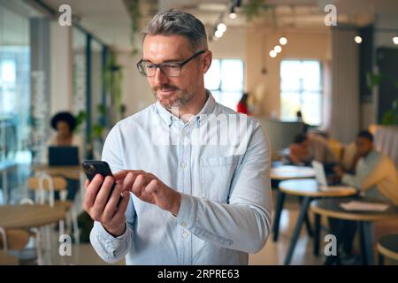 Reifer Lächelnder Geschäftsmann, Der Im Geschäftigen Büro Auf Dem Mobiltelefon Steht Stockfoto