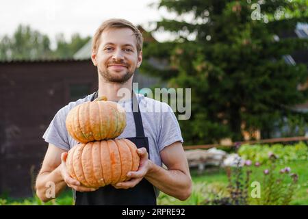 Der junge lächelnde Gärtner in der Schürze hält Kürbisse in seinem Garten. Großartiges Erntekonzept Stockfoto