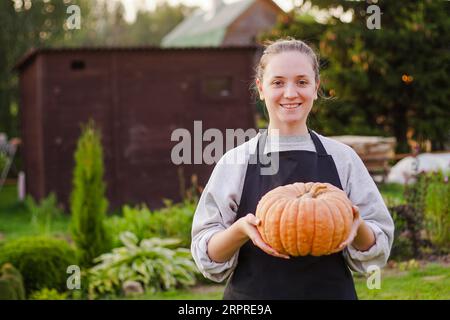 Die junge lächelnde Gärtnerin in in der Schürze hält Kürbisse in seinem Garten. Großartiges Erntekonzept Stockfoto