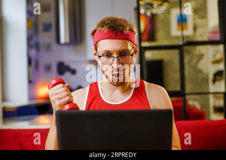 Lustiger junger Mann-Geek mit Kurzhanteln, der online das Trainingsprogramm auf dem Laptop ansieht. Sport- und Humorkonzept. Stockfoto