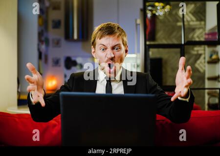 Frustrierter junger Mann im Anzug, der auf der Couch mit pc-Laptop sitzt und schreit. Gestresster Geschäftsmann im Home Office. Stockfoto