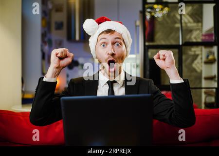 Junger Geschäftsmann in rotem weihnachtsmann Hut sehr glücklich und freut sich vor Laptop zu sitzen. Weihnachtsverkauf, Konzept zum zweiten Weihnachtsfeiertag. Stockfoto
