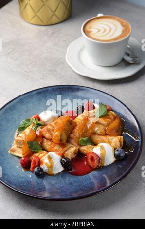 Crepes mit frischen Beeren, karamellisierten Bananen und Toppings, gerollte dünne Pfannkuchen mit Hüttenkäsefüllung auf einem Teller Stockfoto