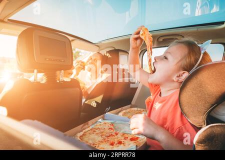 Kleines Mädchen mit offenem Mund Porträt isst italienische Pizza und sitzt im modernen Auto mit Mutter und Vater. Glückliche Familienmomente, Kindheit, Fast Food Stockfoto