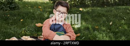 Fröhlicher Schuljunge mit Down-Syndrom, in Brille, Rucksack auf Decke im Park haltend, Banner Stockfoto