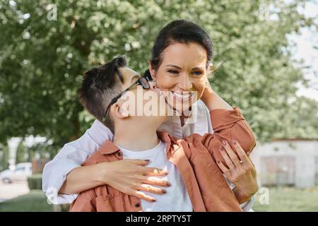 Kind mit Down-Syndrom, das eine lächelnde Mutter mittleren Alters während eines Parkausflugs küsst, glückliche Momente Stockfoto