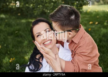 Junge mit Down-Syndrom, in Brille, küssende überfreuliche Mutter im Park, bedingungslose Liebe Stockfoto