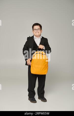 Fröhlicher Schuljunge mit Down-Syndrom, in schwarzem Anzug und Brille mit gelbem Rucksack auf Grau Stockfoto