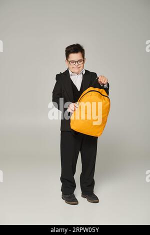 Glücklicher Schuljunge mit Down-Syndrom, in schwarzer Uniform und Brille mit gelbem Rucksack auf Grau Stockfoto