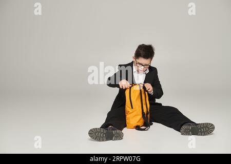 Schuljunge mit Down-Syndrom, in schwarzem Anzug und Brille sitzend mit gelbem Rucksack auf Grau Stockfoto