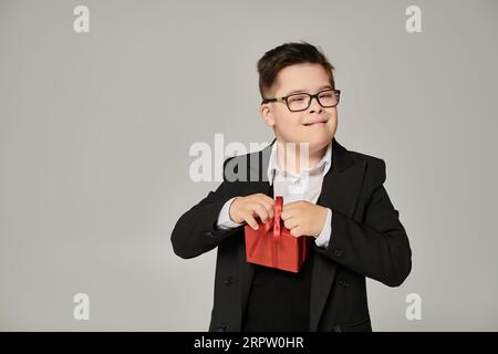 Kind mit Down-Syndrom in Brillen und Schuluniform mit Geschenkbox auf grauem Sonderschüler Stockfoto