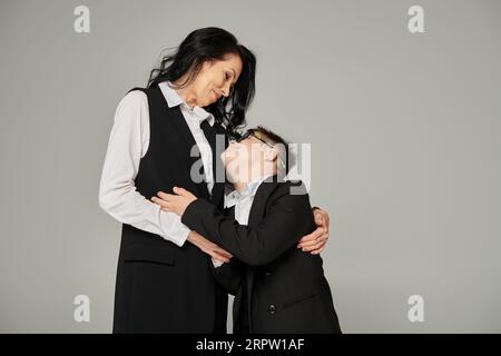 Glückliche Geschäftsfrau, die ein Kind mit Down-Syndrom in Schuluniform und Brille auf Grau umarmt Stockfoto