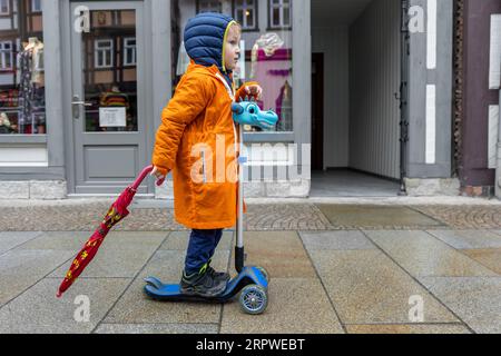 Niedlicher kleiner einsamer kaukasischer Junge in leuchtend orangefarbenem, wasserdichtem Regenmantel. Genießen Sie es, mit dem Roller auf der europäischen Stadtstraße draußen zu spazieren. Herbstwetter Stockfoto