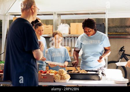 Gruppe von Freiwilligen im Gemeindezentrum, die kostenlose Nahrung an bedürftige Menschen verteilen. Eine afroamerikanische Frau bereitet eine Mahlzeit zu, die einem hungrigen Obdachlosen beim Essen serviert wird. Stockfoto