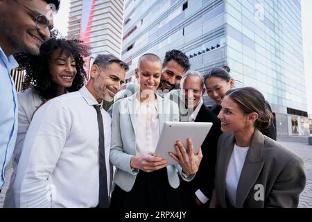 Ein Team aus verschiedenen Profis schaut sich das Tablet von Teamleader an. Fröhliche Kollegen mit vielen verschiedenen Rassen im Freien. Stockfoto