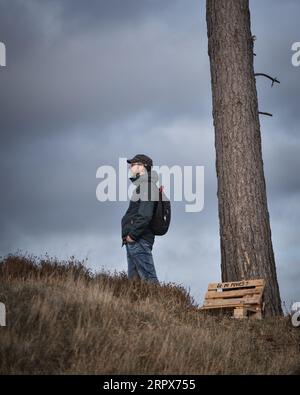 Profilansicht eines Wandermanns mit Händen in der Tasche, der Kappe und Rucksack trägt, vor dunklen Wolken im Hintergrund Stockfoto