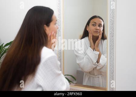 Attraktive Frau im Kleid, die im Spiegel schaut, die Dame, die den Zustand der Gesichtshaut am Frisiertisch überprüft Stockfoto
