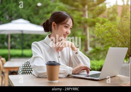 Eine attraktive asiatische Freiberuflerin arbeitet aus der Ferne in einem schönen Café im Freien und nutzt ihren Laptop, um ihre Aufgaben zu verwalten. U Stockfoto