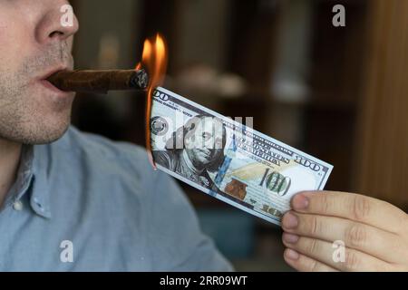 Junger brutaler Mann, der eine Zigarre mit 100 Dollar als Symbol für Reichtum und Erfolg anzündet Stockfoto