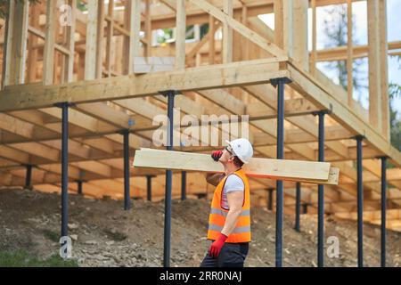 Männlicher Baumeister, der Uniform und Helm trägt, Holzbretter hält, Gilder trägt, nach oben schaut. Stockfoto