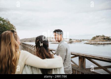 Lächelnder junger Mann mit weiblichen Freunden, die auf der Brücke in der Nähe des Sees spazieren gehen Stockfoto