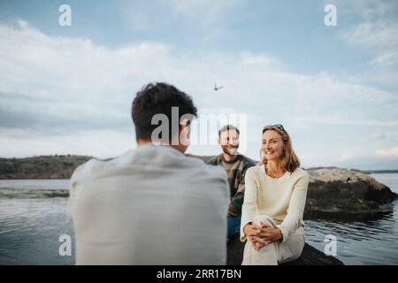 Lächelnde männliche und weibliche Freunde, die miteinander reden, während sie in der Nähe des Sees sitzen Stockfoto