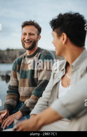 Glücklicher junger Mann, der mit einem Freund in der Nähe des Sees sitzt Stockfoto