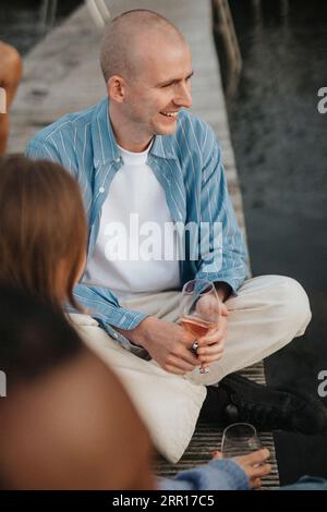 Lächelnder Mann, der Weinglas hält, während er mit den Beinen auf dem Pier sitzt Stockfoto