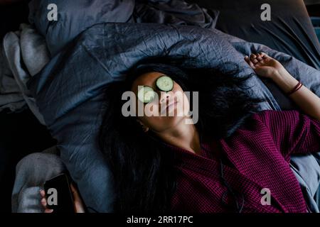 Hochwinkelansicht einer auf dem Bett liegenden Frau mit Gurkenscheiben auf den Augen Stockfoto