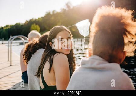 Porträt eines lächelnden Mädchens, das mit Freunden auf dem Steg am See sitzt Stockfoto