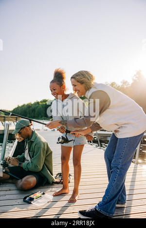 Glückliche Beraterin beim Angeln mit einem Mädchen, das auf dem Steg am See im Sommercamp steht Stockfoto