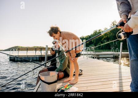 Neugieriger Junge, der im See mit einem Berater beim Angeln auf dem Bootssteg im Sommercamp schaut Stockfoto