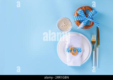 Minimale Tischlage auf dem Oktoberfest. Teller, Gabel, Messer, mit festlich dekorierter Serviette, kariertem Band, Brezel, Bierglas auf blauem Hintergrund. Oktobe Stockfoto