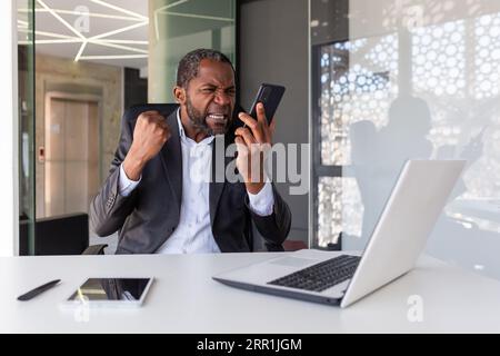 Der ärgerliche, irritierte Boss ruft am Telefon, der Geschäftsmann, der mit dem Gespräch unzufrieden ist, sitzt im Büro, der Mann am Arbeitsplatz in einem Business-Anzug arbeitet mit einem Laptop. Stockfoto