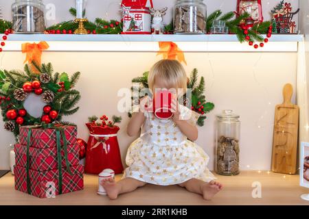 Weihnachtsküche Mädchen. Porträt eines bezaubernden kleinen Mädchens, 2 Jahre alt, sie sitzt auf einem Tisch in einem weißen Kleid und trinkt aus einer roten Tasse in einem dekorierten Stockfoto