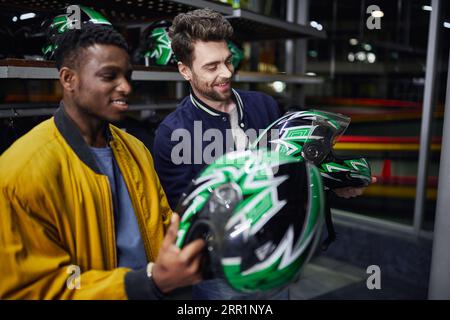 Zwei multikulturelle Freunde in Bomberjacken, die Helme im Inneren der Kartbahn betrachten, Go-Cart Stockfoto