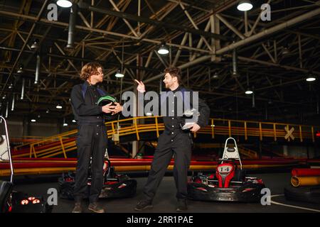 kart Racer zeigt auf einen Freund in der Brille und steht in der Nähe von Rennwagen, während er Helme hält Stockfoto