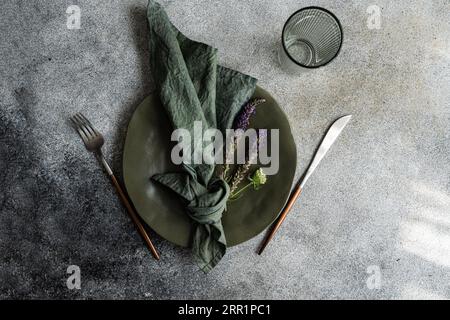 Draufsicht auf herbstliche Tischplatte mit Marmorplatte, Serviette, Gabel und Messer und Lavendelblüten in der Nähe von leerem Glas gegen graue Oberfläche Stockfoto