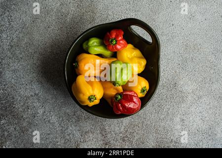 Draufsicht auf verschiedenfarbige Paprika, die in einer runden schwarzen Keramikschale auf einem Betonhintergrund platziert sind Stockfoto