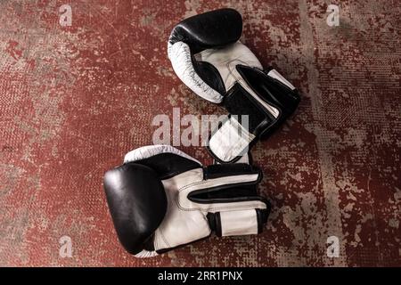 Draufsicht auf ein Paar schwarz-weiß gefärbte Leder-Boxhandschuhe, die auf einem alten roten Boden mit erodierter Oberfläche bei Tageslicht während der Pause in der Praxis platziert werden Stockfoto