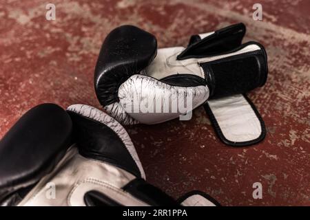 Ein Paar schwarz-weiß gefärbte Boxhandschuhe aus Leder mit hohem Winkel auf einem alten roten Boden mit erodierter Oberfläche bei Tageslicht während der Pause in der Praxis Stockfoto