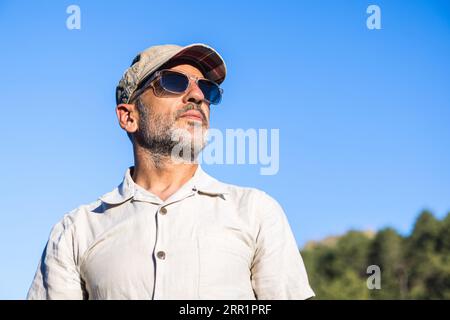 Ein kleiner, selbstbewusster bärtiger Mann, der ein weißes Hemd und eine Sonnenbrille trägt, während er unter blauem Himmel steht Stockfoto