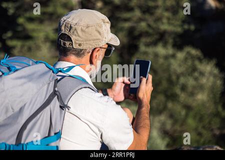 Seitenansicht des anonymen bärtigen männlichen Touristen in der Kappe mit Rucksack, der am Rand der felsigen Klippe über grünen Pflanzen sitzt und das Handy durchstöbert Stockfoto