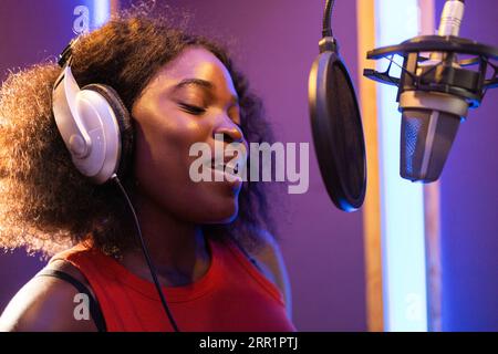 Seitenansicht einer afroamerikanischen professionellen Sängerin in Kopfhörern, die beim Singen und Aufnehmen in musikalischen Studien in der Nähe des Mikrofons hohe Noten macht Stockfoto