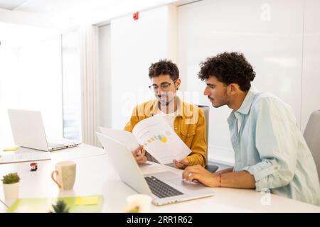 Männliche Kollegen sitzen am weißen Schreibtisch im modernen Büro und arbeiten gemeinsam mit Laptop und Papieren an Projekten Stockfoto