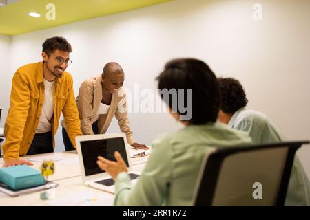 Ein lächelnder junger Mann in einer Brille und eine anonyme Frau am Tisch mit verschiedenen Mitgliedern, die sich beim Projektgespräch anschauen Stockfoto
