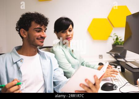 Lächelnde junge, multirassische Kollegen, die nach unten schauen, während sie am Tisch sitzen, mit Notizen und einem Stift, der hispanischen Mann zeigt, dass Frauen auf der Tastatur arbeiten Stockfoto